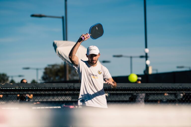 Player in a white cap raising his paddle high to hit an overhead near the net