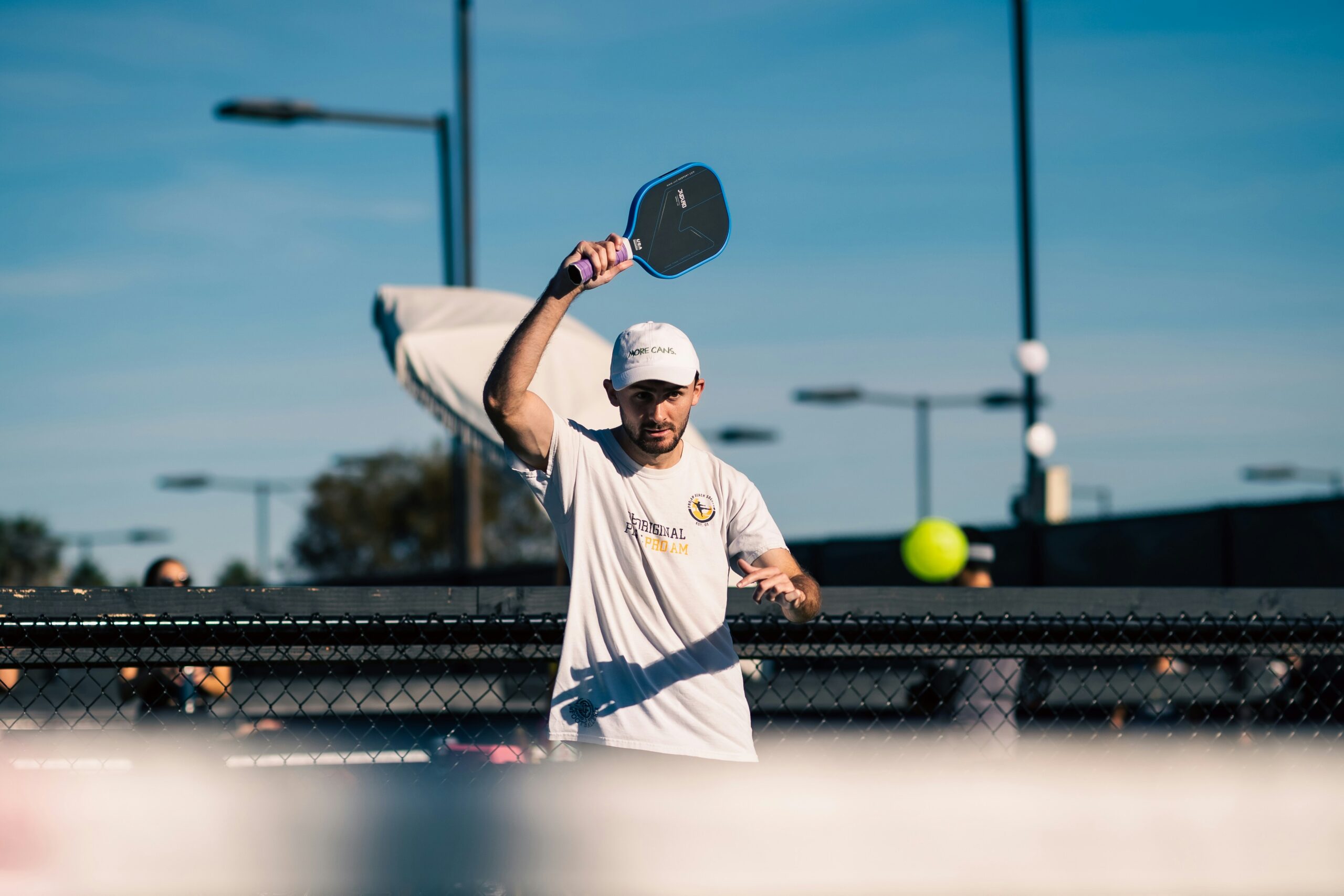 Player in a white cap raising his paddle high to hit an overhead near the net