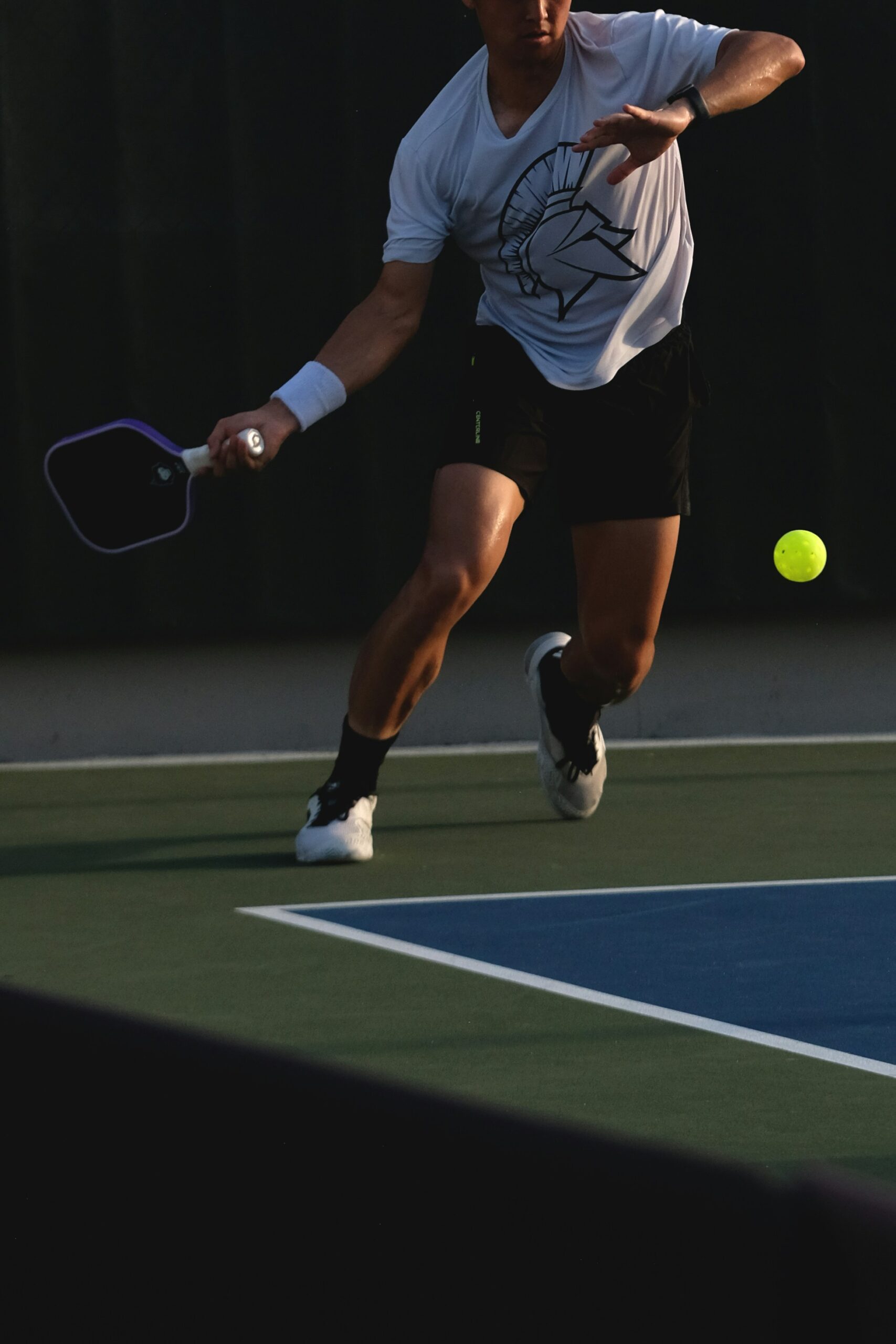 Player lunging forward to reach a low ball near the kitchen line, lit against a dark background
